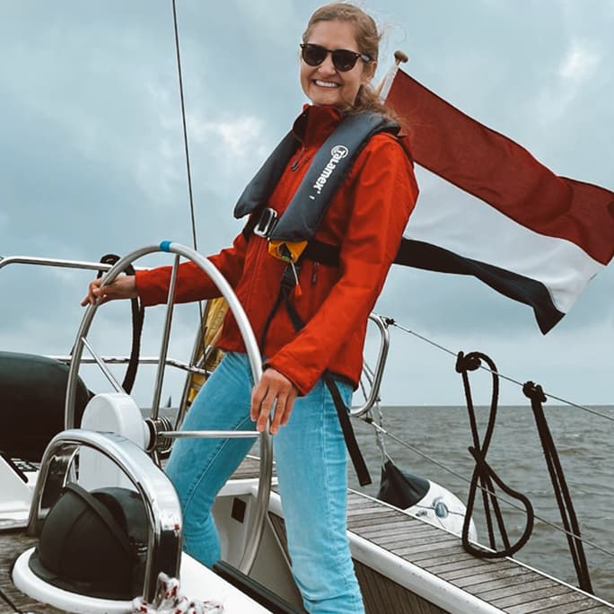 Smiling sailor in a red jacket and life vest at the helm of a yacht with a Dutch flag flying behind