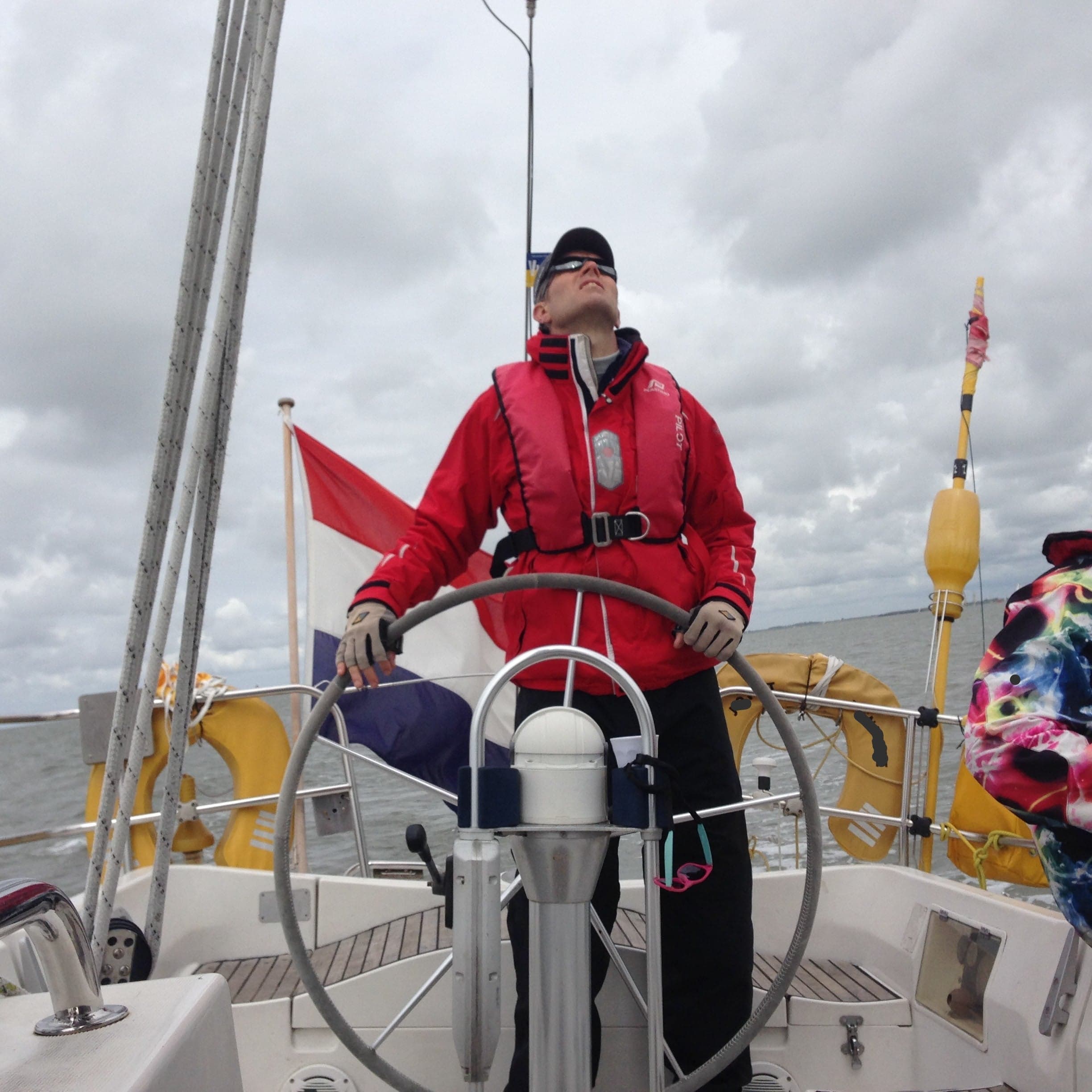 Skipper in a red lifejacket at the helm of a yacht looking up at the rig under grey skies