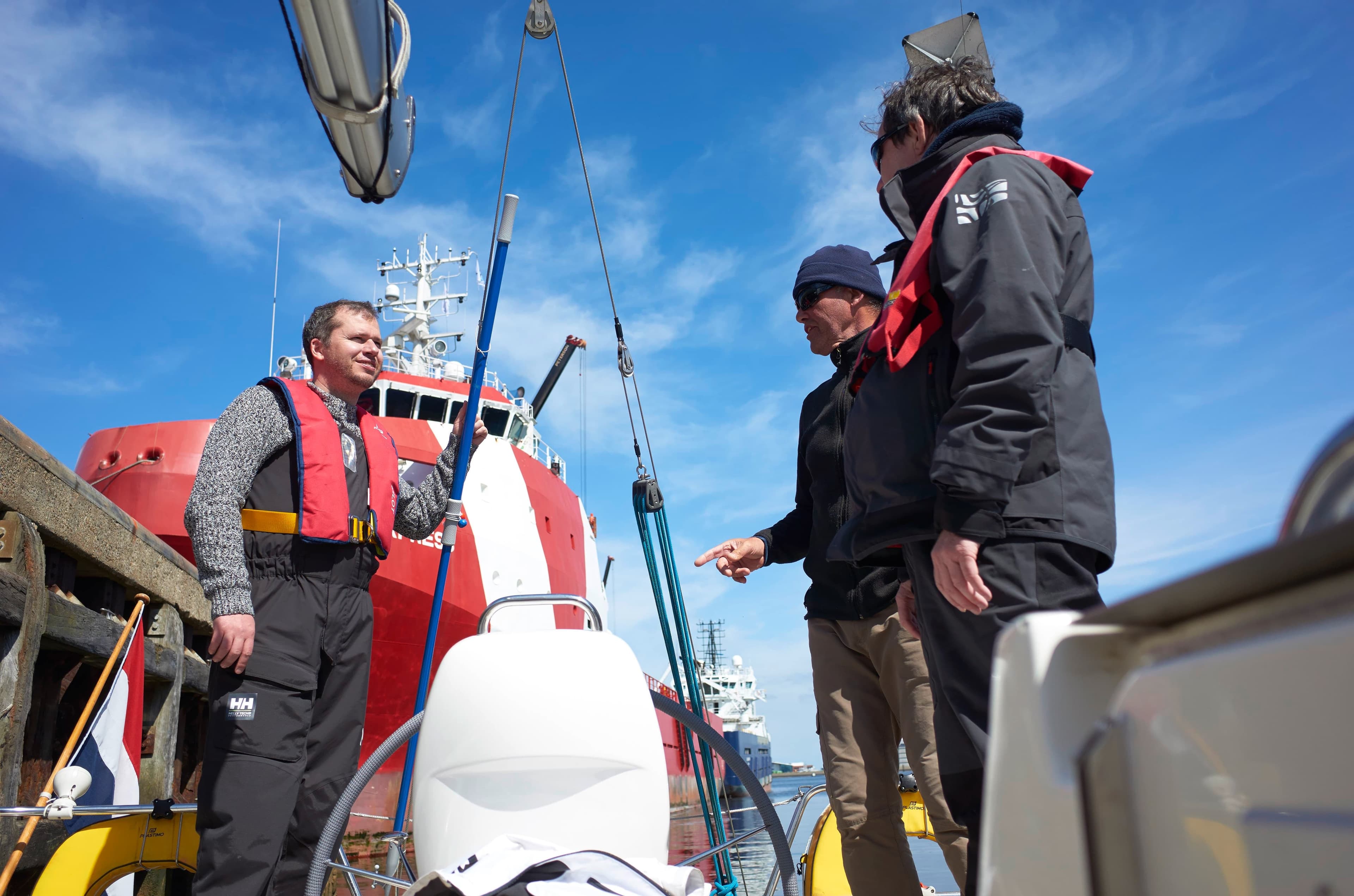 Three sailors in conversation on deck of a yacht with a red coastguard vessel docked behind them